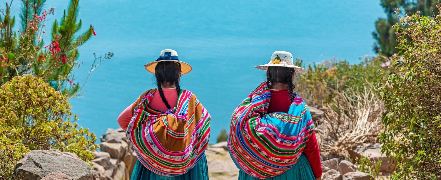Two indigenous Quechua women in traditional clothes walking down the path to the harbor of Isla Taquile (Taquile Island) with the Titicaca Lake in the background | Peru | Swift Travel